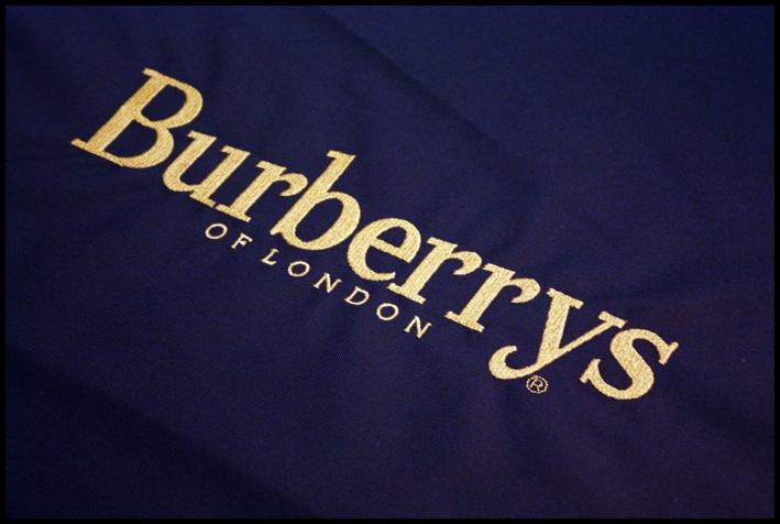 Granatowa tkanina z wyhaftowanym złotą nicią napisem 'Burberrys OF LONDON' pod kątem, ukazująca detale ściegów.