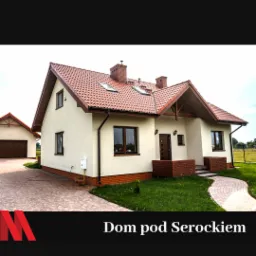 Gotowy dom jednorodzinny z beżową elewacją, czerwoną dachówką i ceglanymi elementami dekoracyjnymi przy wejściu, z dodatkowym budynkiem gospodarczym w tle.