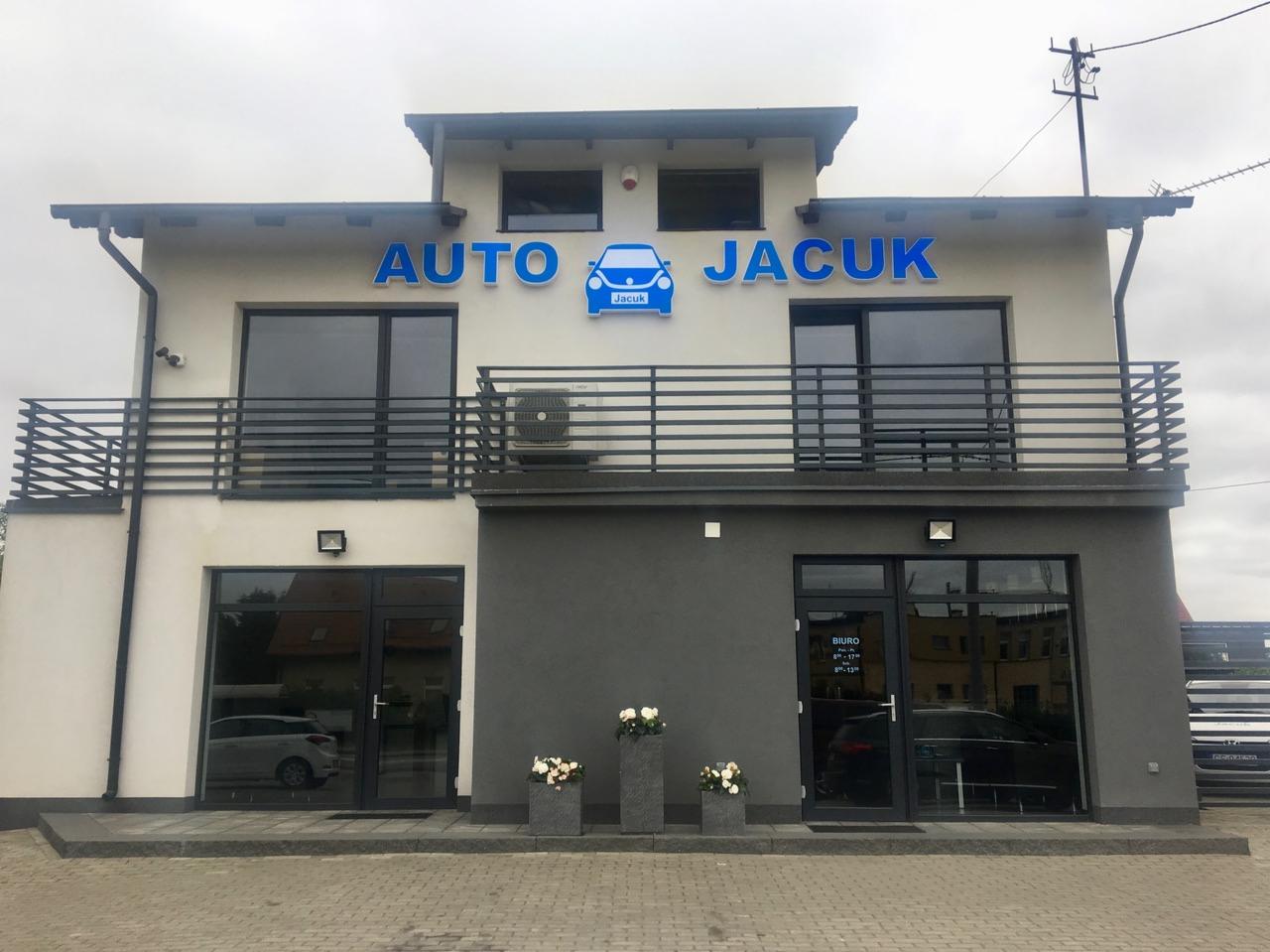 Budynek warsztatu samochodowego z niebieskim szyldem 'Auto Jacuk', balkonem i trzema donicami z kwiatami przed wejściem.