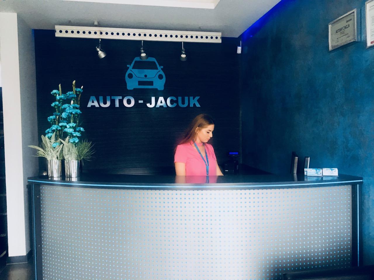 Recepcja warsztatu samochodowego z logo Auto-Jacuk, kwiatami i pracownicą w różowej koszuli.