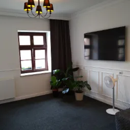 Pok&oacute;j z ciemną wykładziną dywanową, białymi ścianami z dekoracyjnymi panelami, brązowym oknem, ciemnymi zasłonami i czarnym żyrandolem.