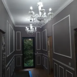 Korytarz w trakcie wykańczania, ściany w kolorze taupe z białymi, dekoracyjnymi listwami, drewnopodobna podłoga, kryształowe żyrandole, widok na zieleń przez drzwi balkonowe.