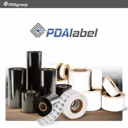 Różne rolki etykiet samoprzylepnych, zarówno czarne termotransferowe, jak i białe, ułożone na brązowym blacie.