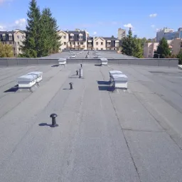 Szary, płaski dach pokryty papą z kominkami wentylacyjnymi i wywietrznikami, na tle budynków mieszkalnych i błękitnego nieba z chmurami.