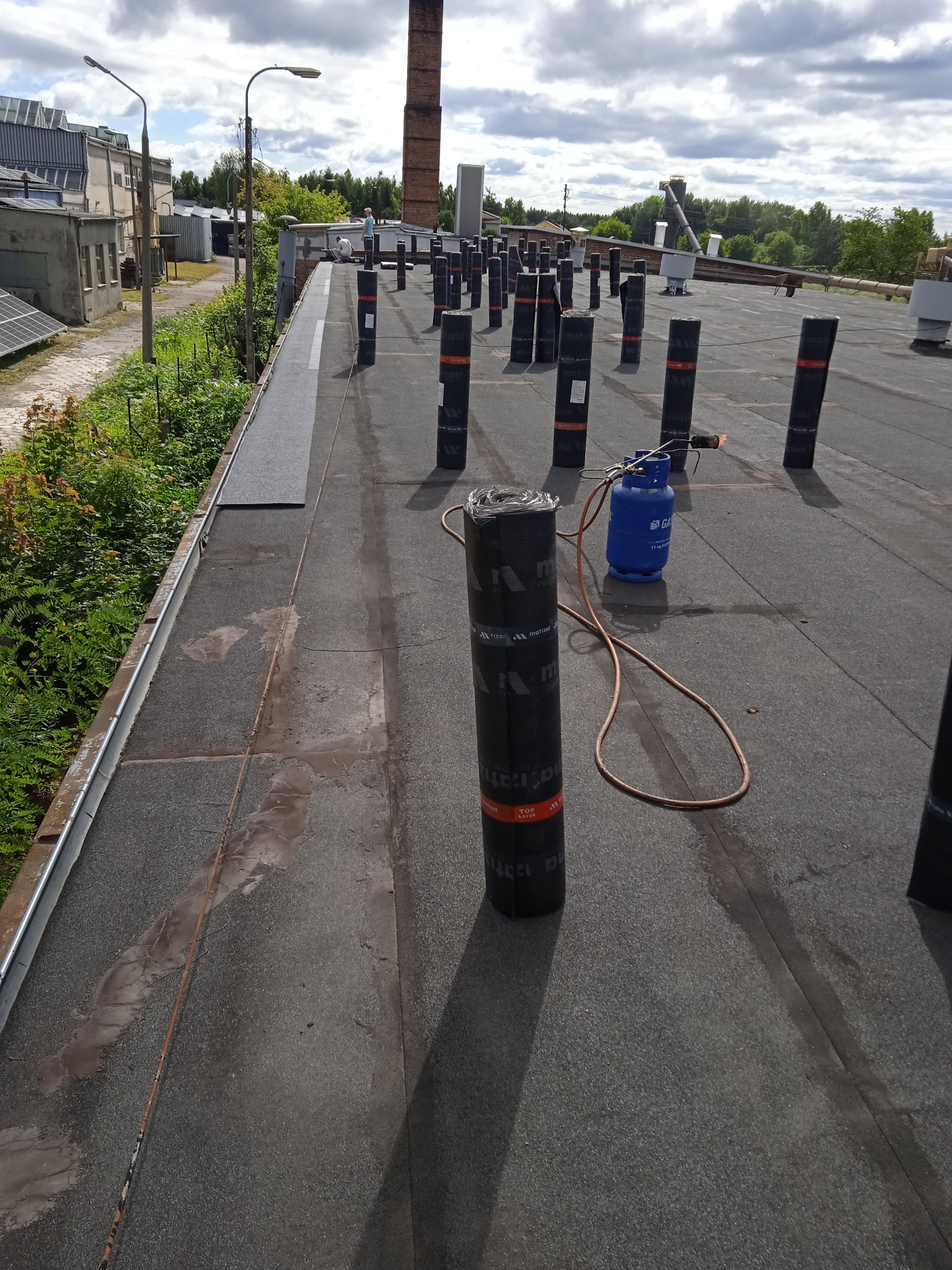 Prace dekarskie na płaskim dachu: rolki papy, butla gazowa i palnik w użyciu. W tle komin i budynki przemysłowe.