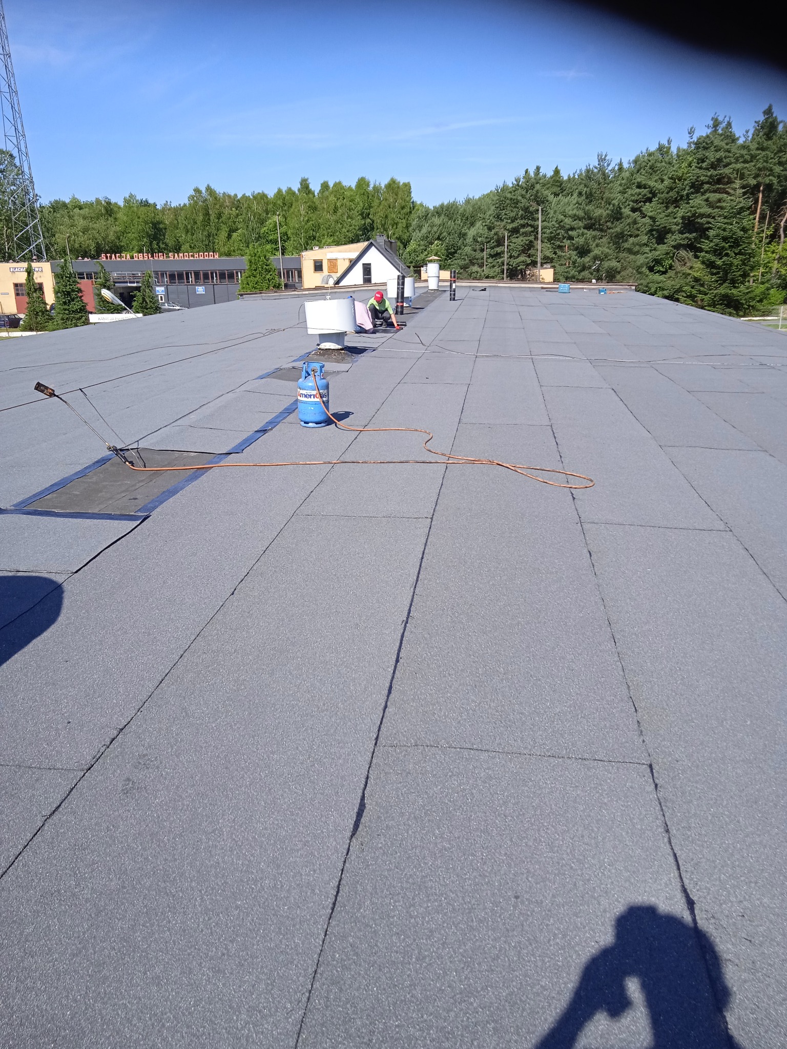 Prace dekarskie na płaskim dachu pokrytym papą termozgrzewalną. Widoczny palnik, butla gazowa i dekarz w oddali. W tle budynek stacji obsługi samochodów.