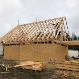 Budowa domu w trakcie realizacji: szkielet drewniany z desek i płyt OSB, widoczni pracownicy na rusztowaniu montujący konstrukcję dachu, na pierwszym planie stos drewna budowlanego.