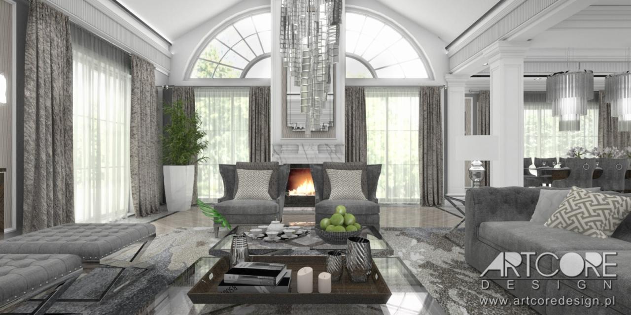 Wnętrze przestronnego salonu z kominkiem, wysokimi oknami łukowymi, szarymi meblami i dekoracyjnym żyrandolem. Na stoliku kawowym książki i świece, w tle widoczna jadalnia.
