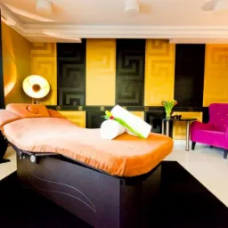 Pomarańczowe łóżko do zabiegów w eleganckim gabinecie kosmetycznym z czarno-złotą ścianą w greckie wzory, zwinięte białe ręczniki, tulipany w wazonie i różowy fotel.