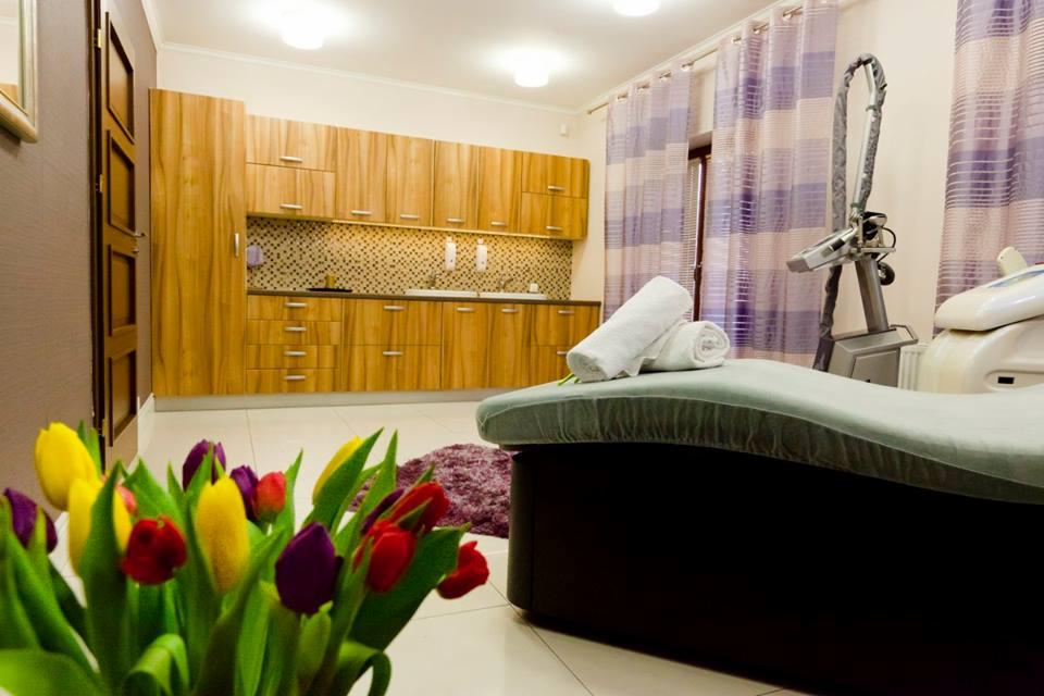 Jasne wnętrze salonu kosmetycznego z łóżkiem do zabiegów, zwiniętymi ręcznikami, urządzeniem kosmetycznym i bukietem tulipanów w pierwszym planie.