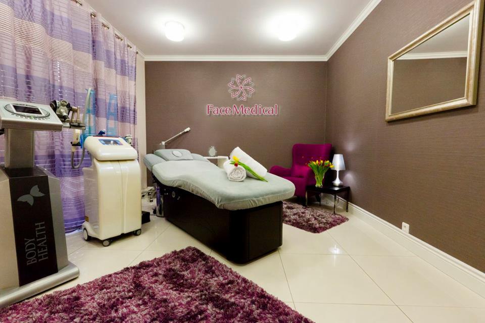 Nowoczesne wnętrze gabinetu z łóżkiem do zabiegów, urządzeniami kosmetycznymi, fotelem i dekoracjami w odcieniach fioletu i brązu, z logo 'Face Medical' na ścianie.
