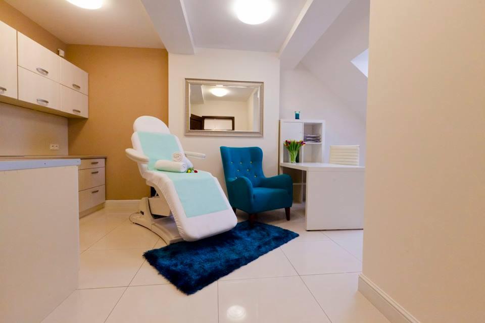 Jasne wnętrze gabinetu z fotelem kosmetycznym z turkusowym obiciem, turkusowym dywanikiem i fotelem, biurkiem z tulipanami i szafkami w tle.