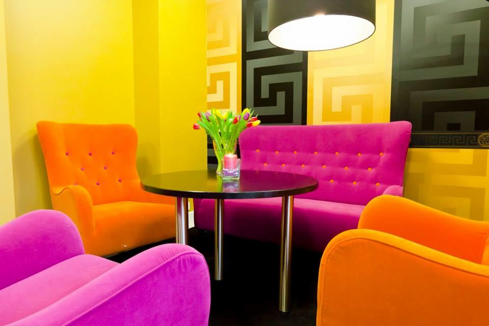 Żywe kolory w poczekalni: fuksjowe i pomarańczowe welurowe fotele wokół czarnego stolika z bukietem tulipanów, na tle żółto-czarnej ściany z geometrycznym wzorem.