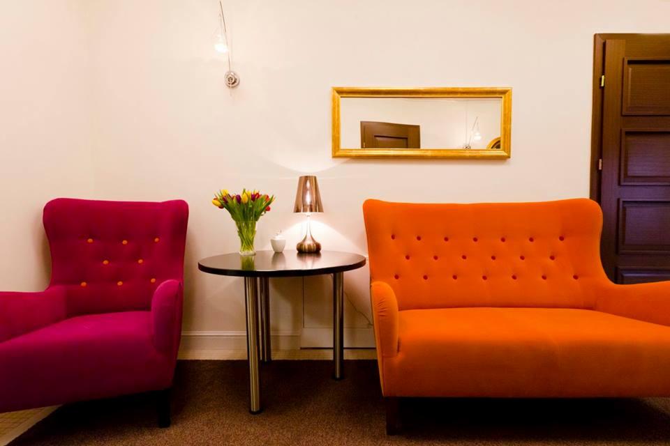 Wnętrze poczekalni z fotelem w kolorze fuksji i pomarańczową sofą, okrągłym stolikiem z tulipanami i lampką, oraz lustrem w złotej ramie na ścianie.