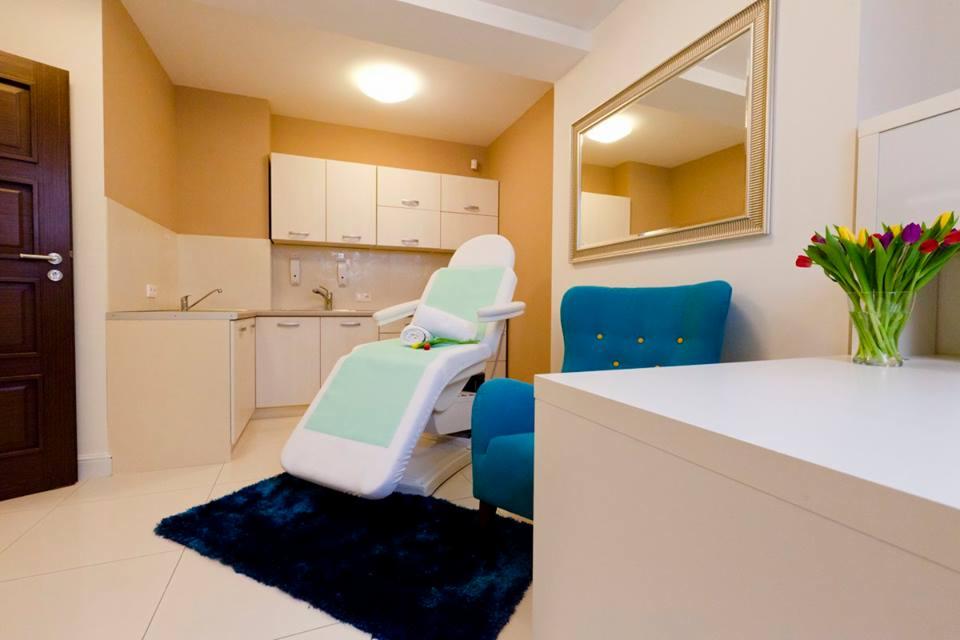 Jasne wnętrze gabinetu z fotelem kosmetycznym z zielonym obiciem, niebieskim dywanikiem i bukietem tulipanów na białym stoliku.