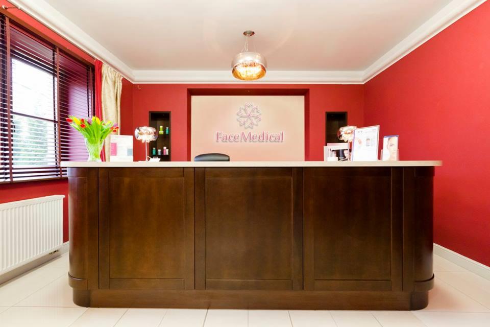 Recepcja z ciemnego drewna w gabinecie kosmetycznym z czerwonymi ścianami, bukiet tulipanów w wazonie na blacie, logo 'Face Medical' na ścianie w tle.