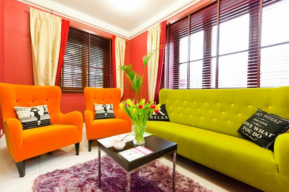 Wnętrze salonu kosmetycznego z pomarańczowymi fotelami, limonkową sofą, stolikiem kawowym z tulipanami i fioletowym dywanem, oświetlone naturalnym światłem z okien z drewnianymi żaluzjami.