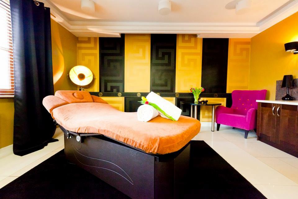 Pomarańczowe łóżko do zabiegów w eleganckim gabinecie kosmetycznym z czarno-złotą ścianą w greckie wzory, zwinięte białe ręczniki, tulipany w wazonie i różowy fotel.