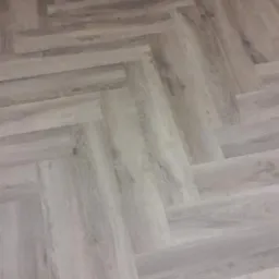 Jasnoszara wykładzina podłogowa ułożona w jodełkę, imitująca drewno, z widoczną strukturą i delikatnym połyskiem.