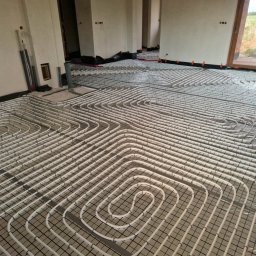 Tomasz Ciężki-Hydro-Tom - Białe rury ogrzewania podłogowego ułożone na czarnej siatce w surowym wnętrzu z dużym oknem. Widoczne elementy instalacji i przygotowanie do wylewki.