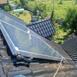 Tomasz Ciężki-Hydro-Tom - Zamontowany kolektor słoneczny na czarnym, błyszczącym dachu z dachówki ceramicznej. W tle zielony ogród z trampoliną i szklarnią w słoneczny dzień.