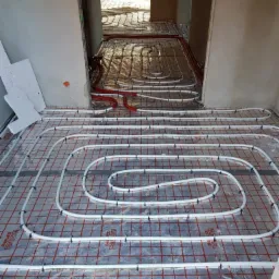 Rozłożona instalacja ogrzewania podłogowego z białych rurek na folii z czerwoną siatką, z widocznymi mocowaniami, w surowym wnętrzu budynku.