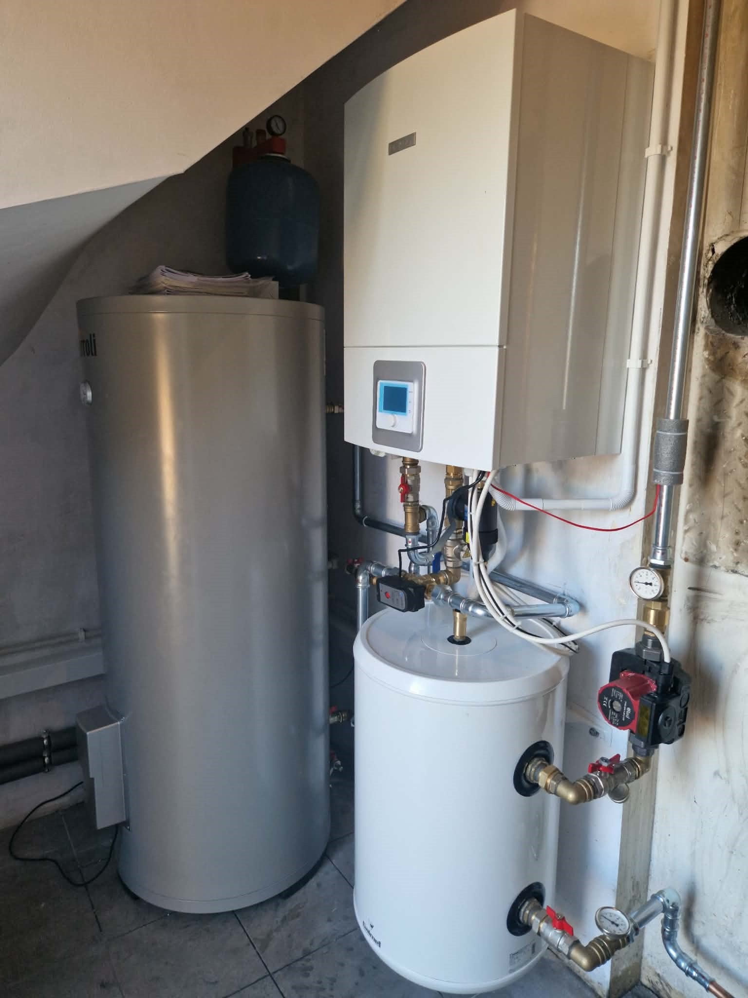 Zestaw urządzeń grzewczych: srebrny zasobnik ciepłej wody, biały bojler i piec gazowy z wyświetlaczem, połączone rurami w kotłowni.