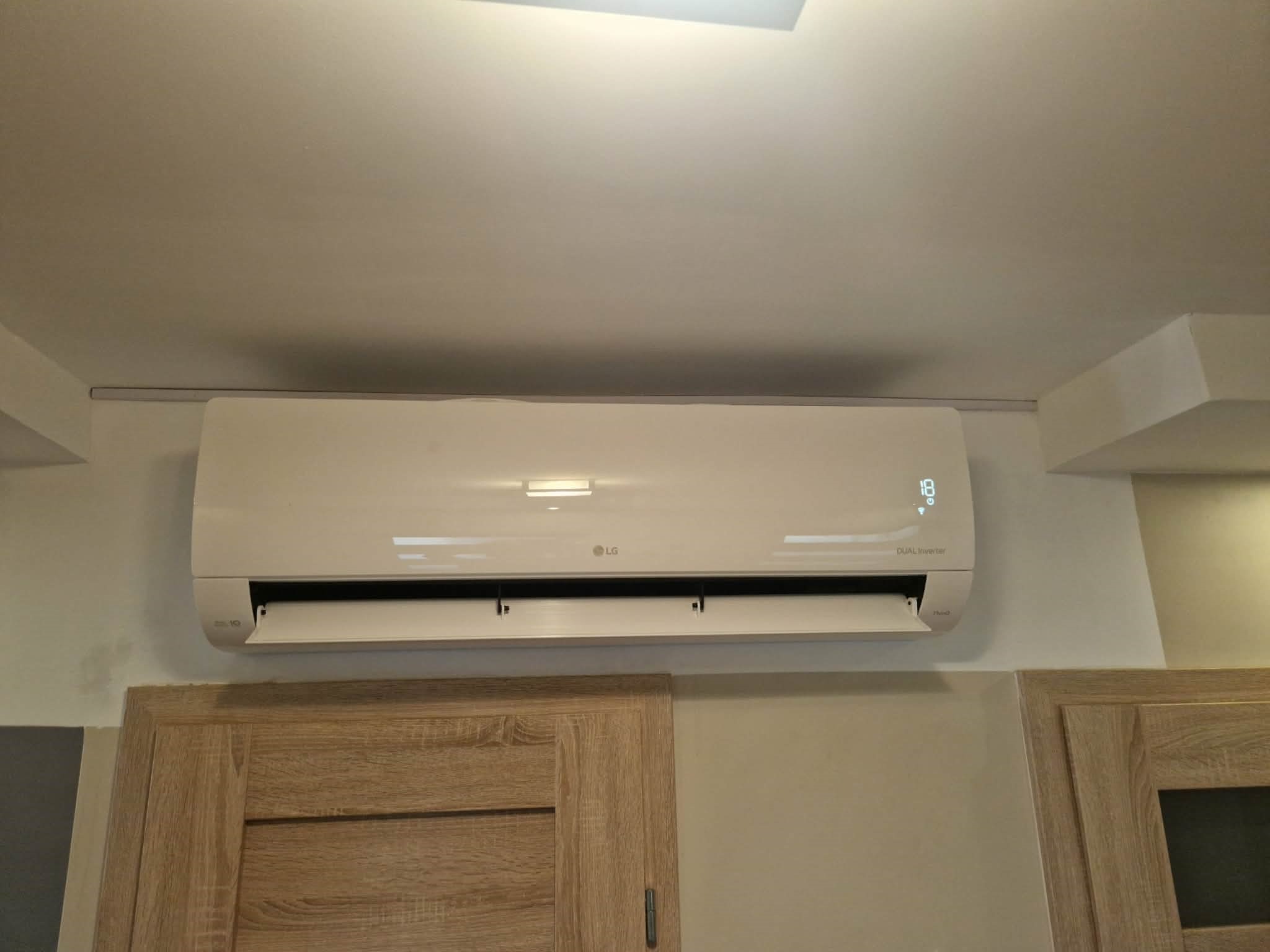 Biały klimatyzator LG Dual Inverter zamontowany na ścianie nad drewnianymi drzwiami szafki, widoczny wyświetlacz temperatury.