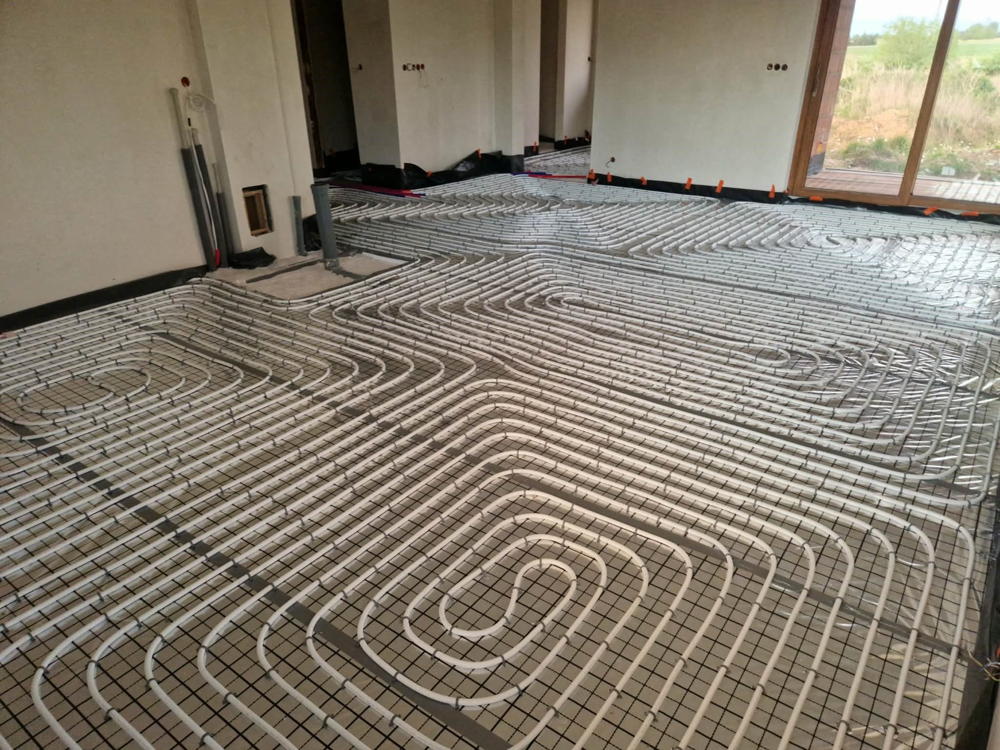 Białe rury ogrzewania podłogowego ułożone na czarnej siatce w surowym wnętrzu z dużym oknem. Widoczne elementy instalacji i przygotowanie do wylewki.