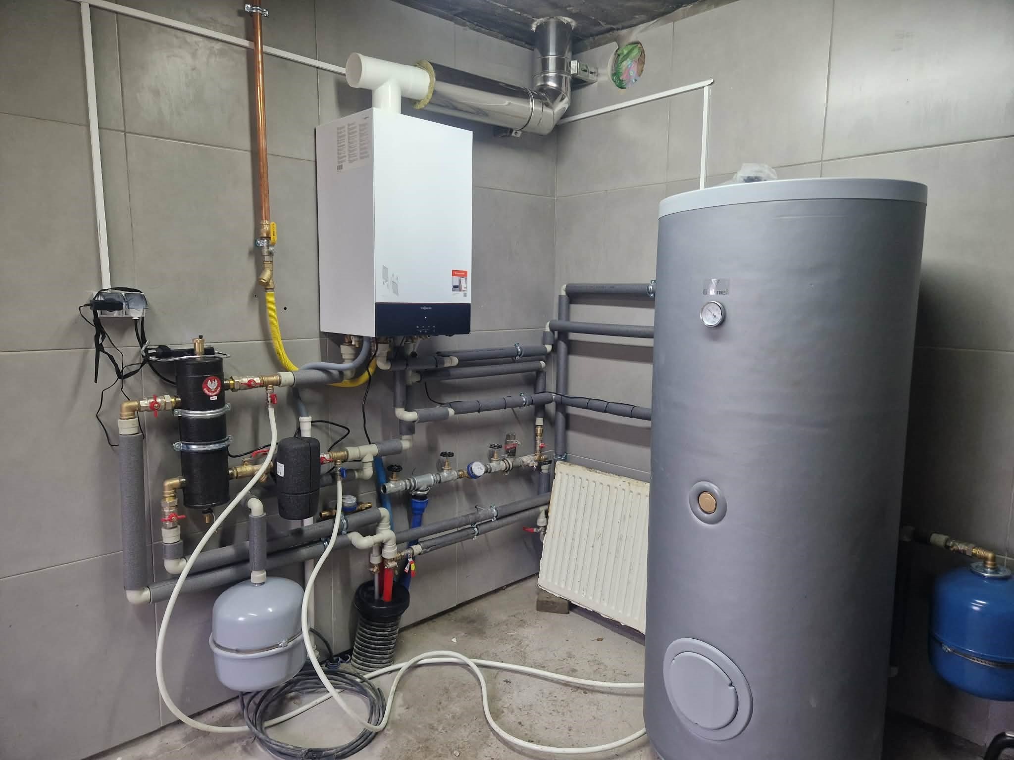 Złożona instalacja grzewcza w kotłowni: piec gazowy, zasobnik ciepłej wody, rury i armatura. Szare płytki na ścianach, betonowy sufit. Widok na kompletną, działającą instalację.