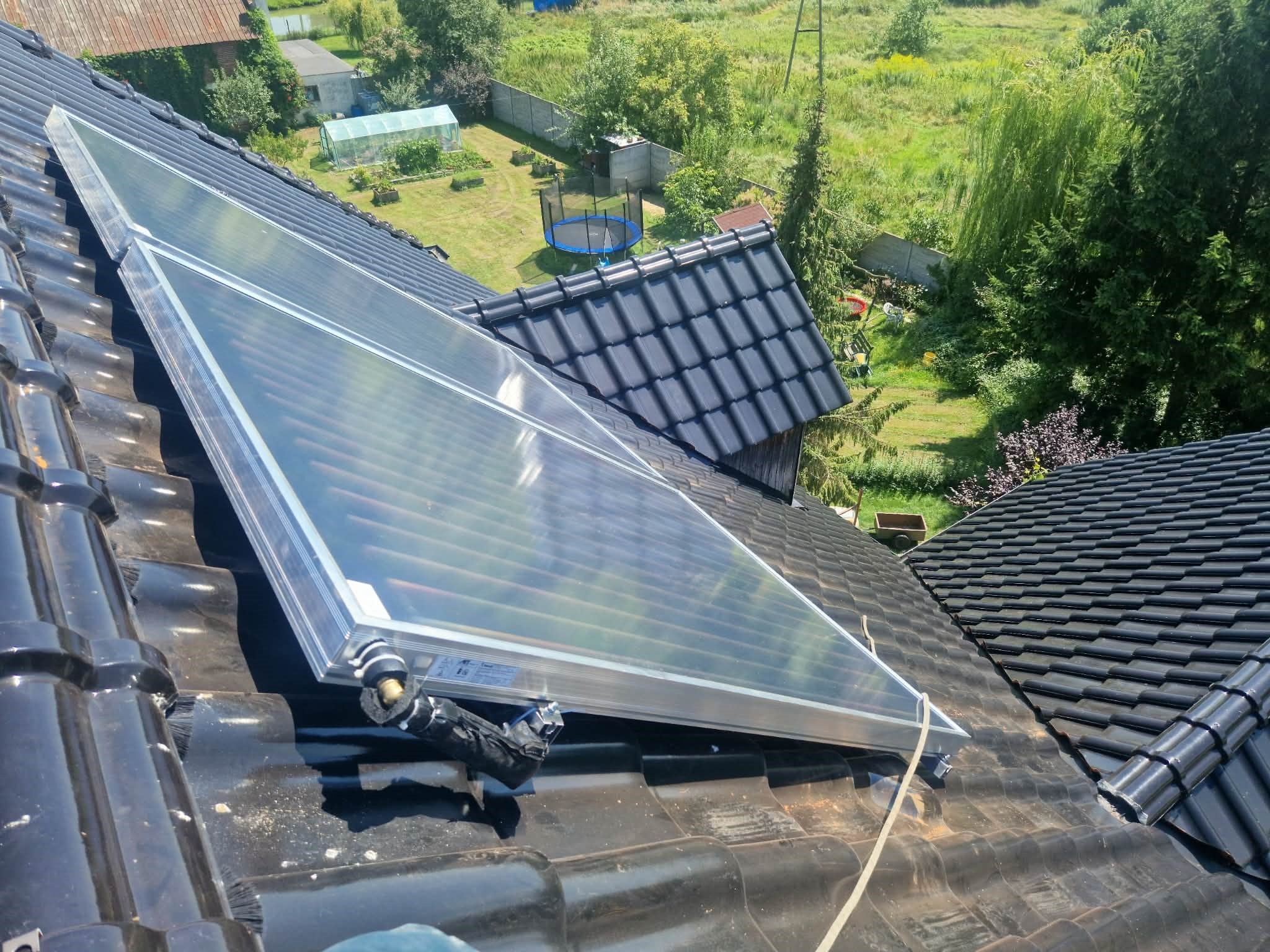 Zamontowany kolektor słoneczny na czarnym, błyszczącym dachu z dachówki ceramicznej. W tle zielony ogród z trampoliną i szklarnią w słoneczny dzień.