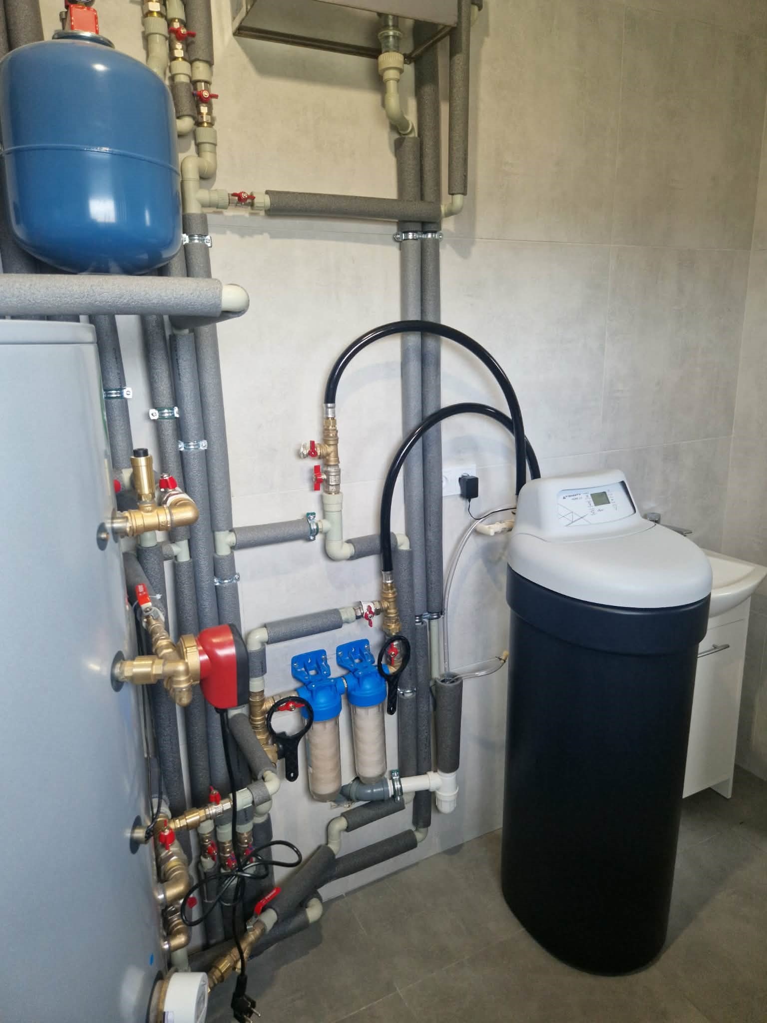 Złożona instalacja hydrauliczna z filtrami, rurami w izolacji i zbiornikiem wyrównawczym w kotłowni z szarą glazurą. Widoczny zmiękczacz wody.