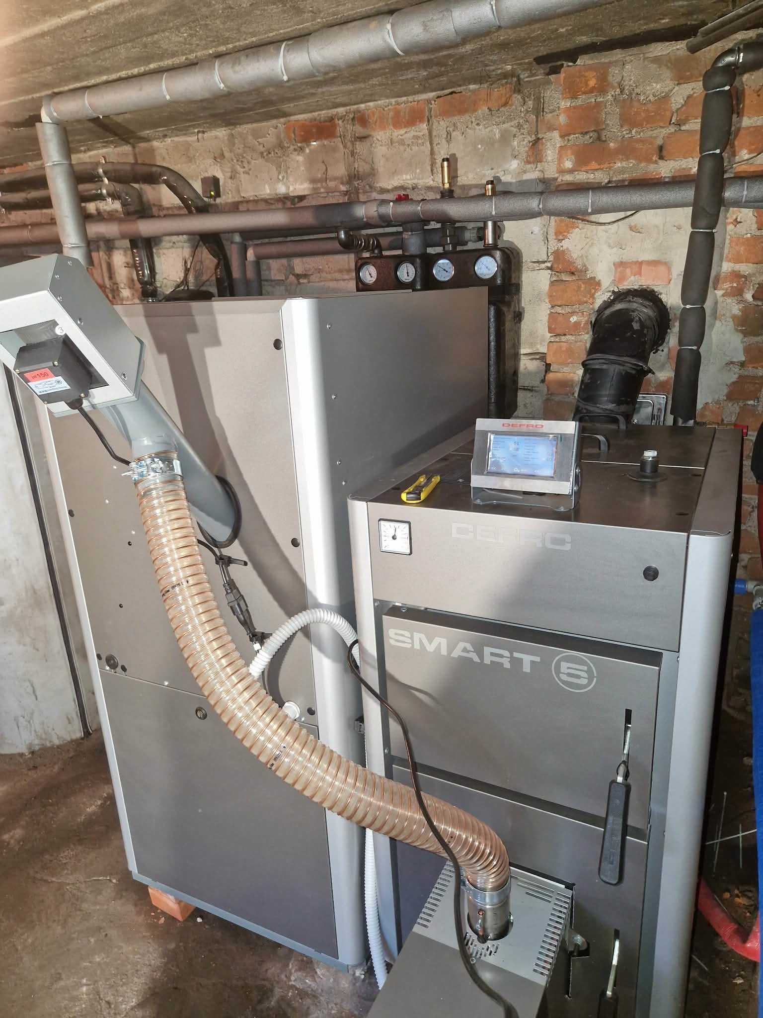 Nowoczesny, srebrny piec Defro Smart E z podajnikiem, widoczne rury instalacji grzewczej na tle ceglanej ściany. Żółty nożyk na piecu.