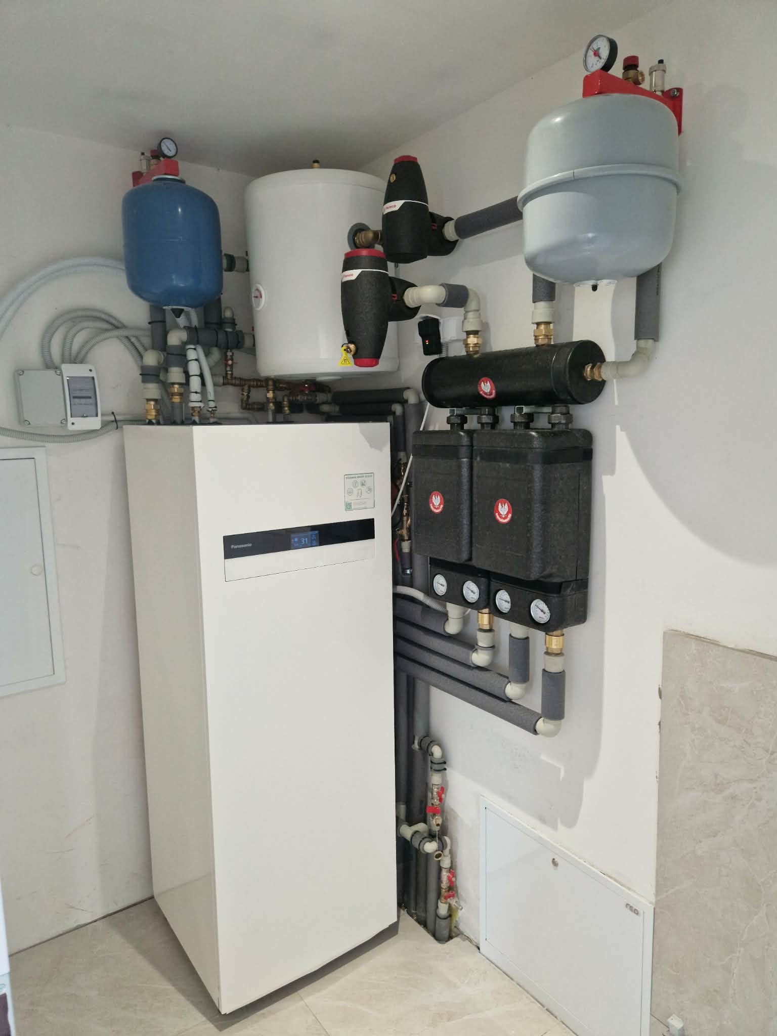 Biała pompa ciepła Panasonic z zbiornikami wyrównawczymi i czarnymi modułami hydraulicznymi na ścianie w jasnym pomieszczeniu. Widoczne rury i manometry.