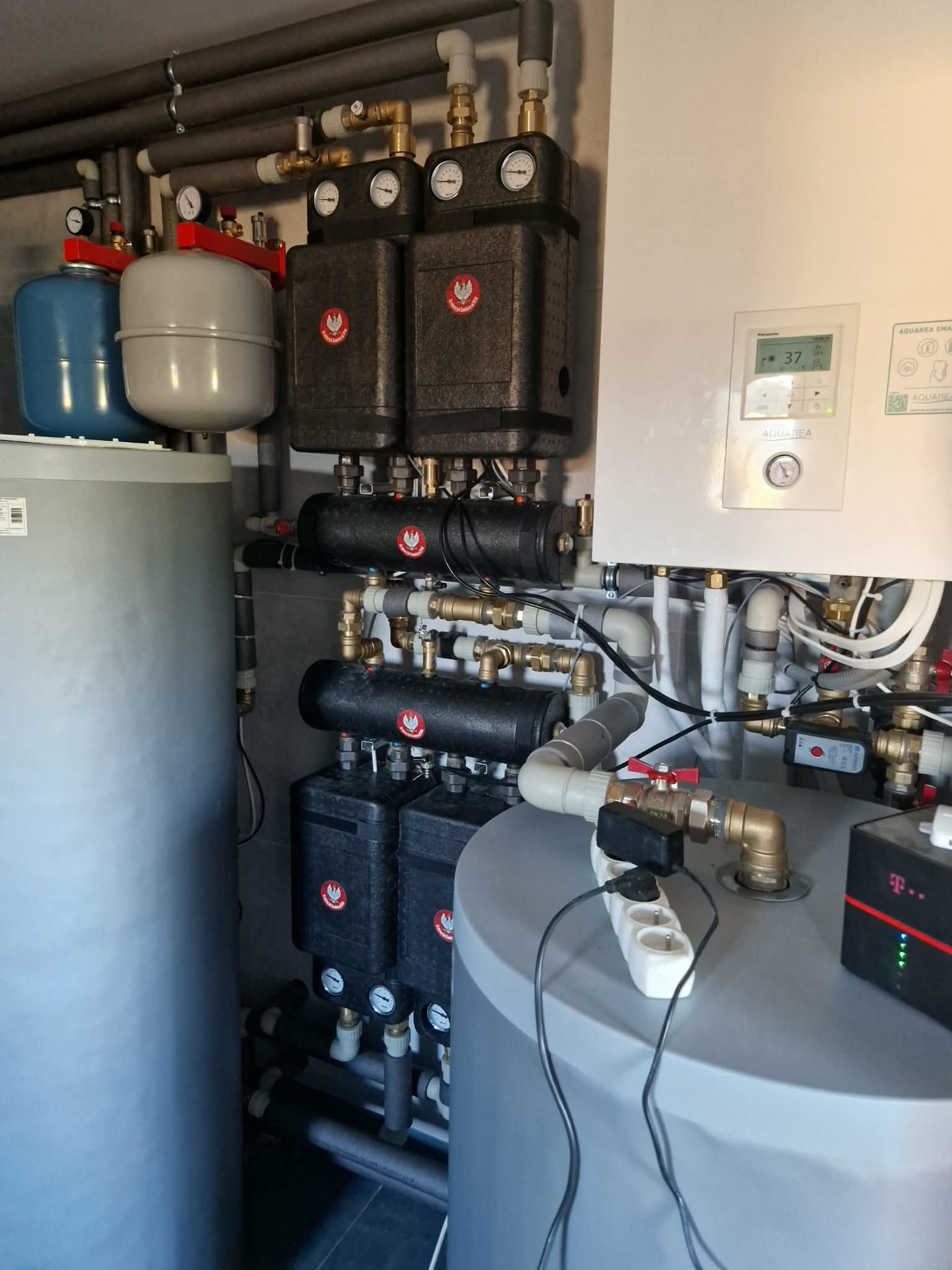 Złożona instalacja grzewcza z zbiornikami, pompami i rurami w pomieszczeniu technicznym. Widoczne manometry i sterownik. Elementy połączone przewodami elektrycznymi.