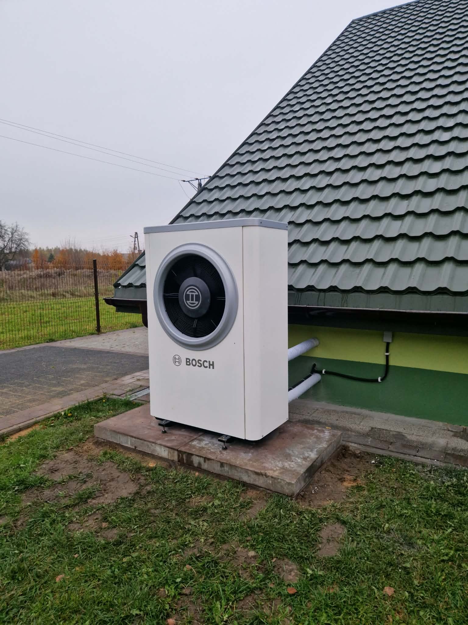 Biała pompa ciepła Bosch na betonowym fundamencie, z widocznymi rurami przy budynku z zielonym dachem i elewacją. Tło: ogrodzenie i drzewa.