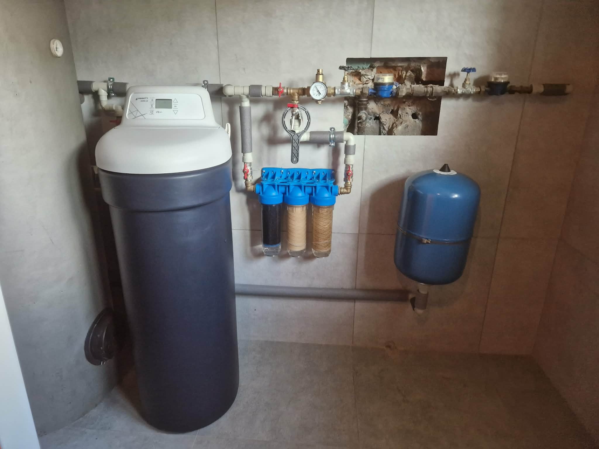 Zestaw filtrów wody z trzema przezroczystymi obudowami, manometrem i niebieskim zbiornikiem ciśnieniowym na tle płytek ceramicznych.