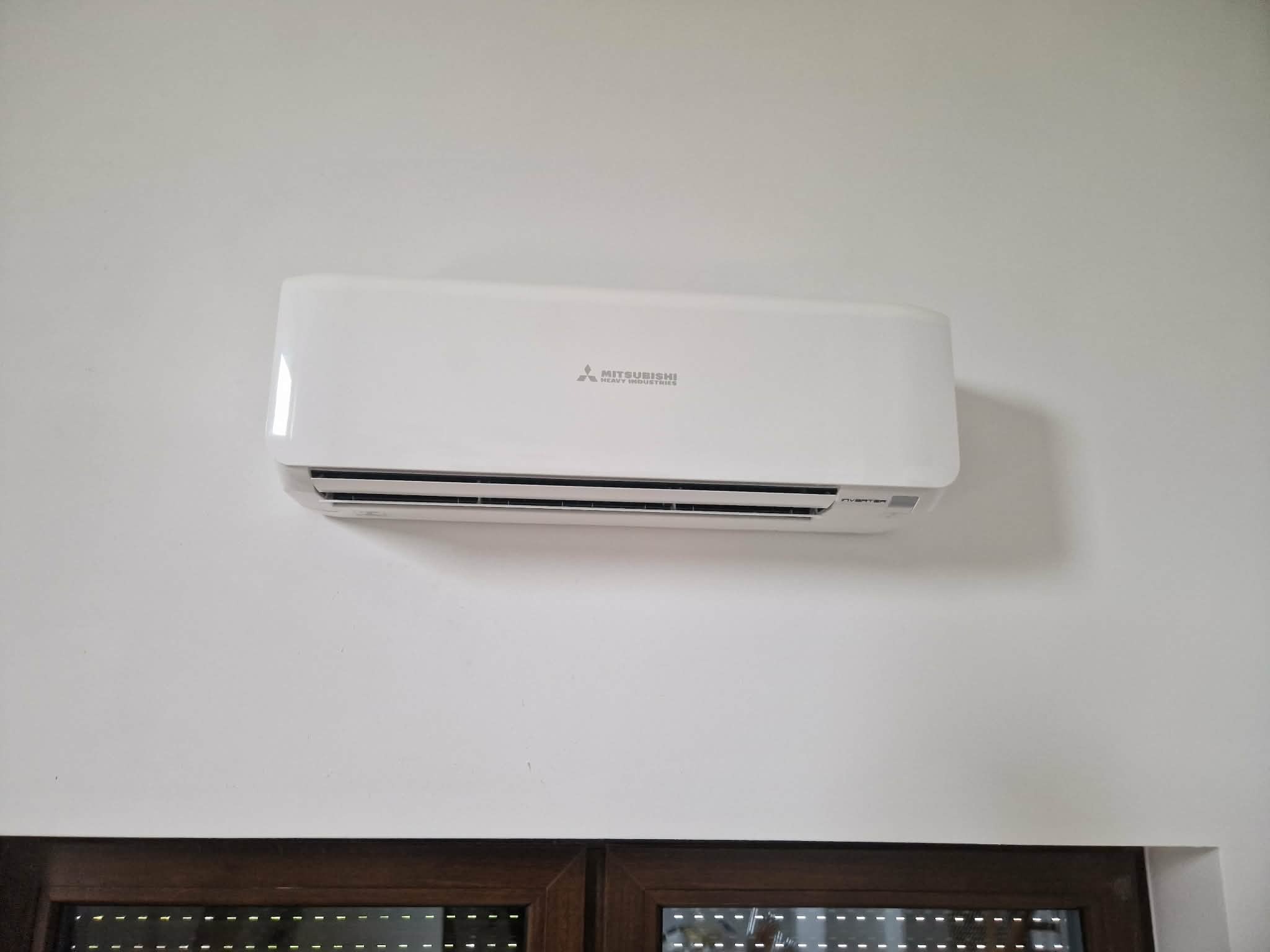 Biały klimatyzator Mitsubishi zamontowany na ścianie nad oknem z brązowymi roletami. Widoczne logo producenta i napis 'Inverter'.