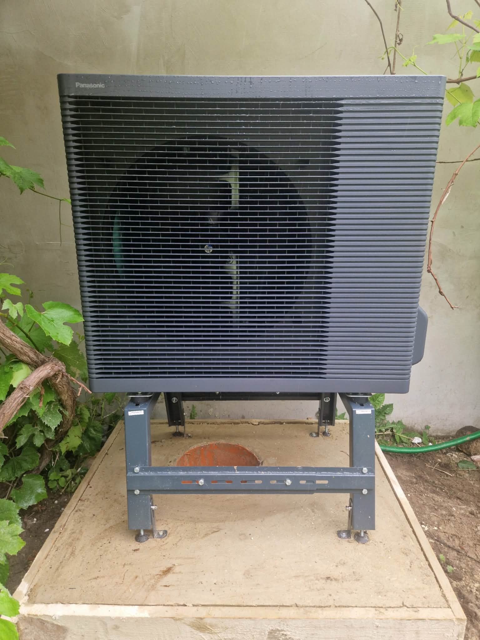 Szara pompa ciepła Panasonic Aquarea zamontowana na metalowej konstrukcji, na betonowej podstawie. W tle ściana i zieleń.