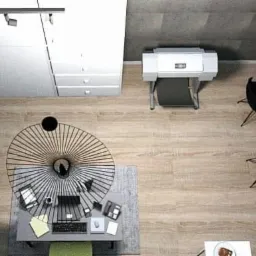 Widok z góry na nowoczesne biuro z drewnianą podłogą, długim biurkiem z laptopami, okrągłym stolikiem z lampą, drukarką i planami na stole w rogu.