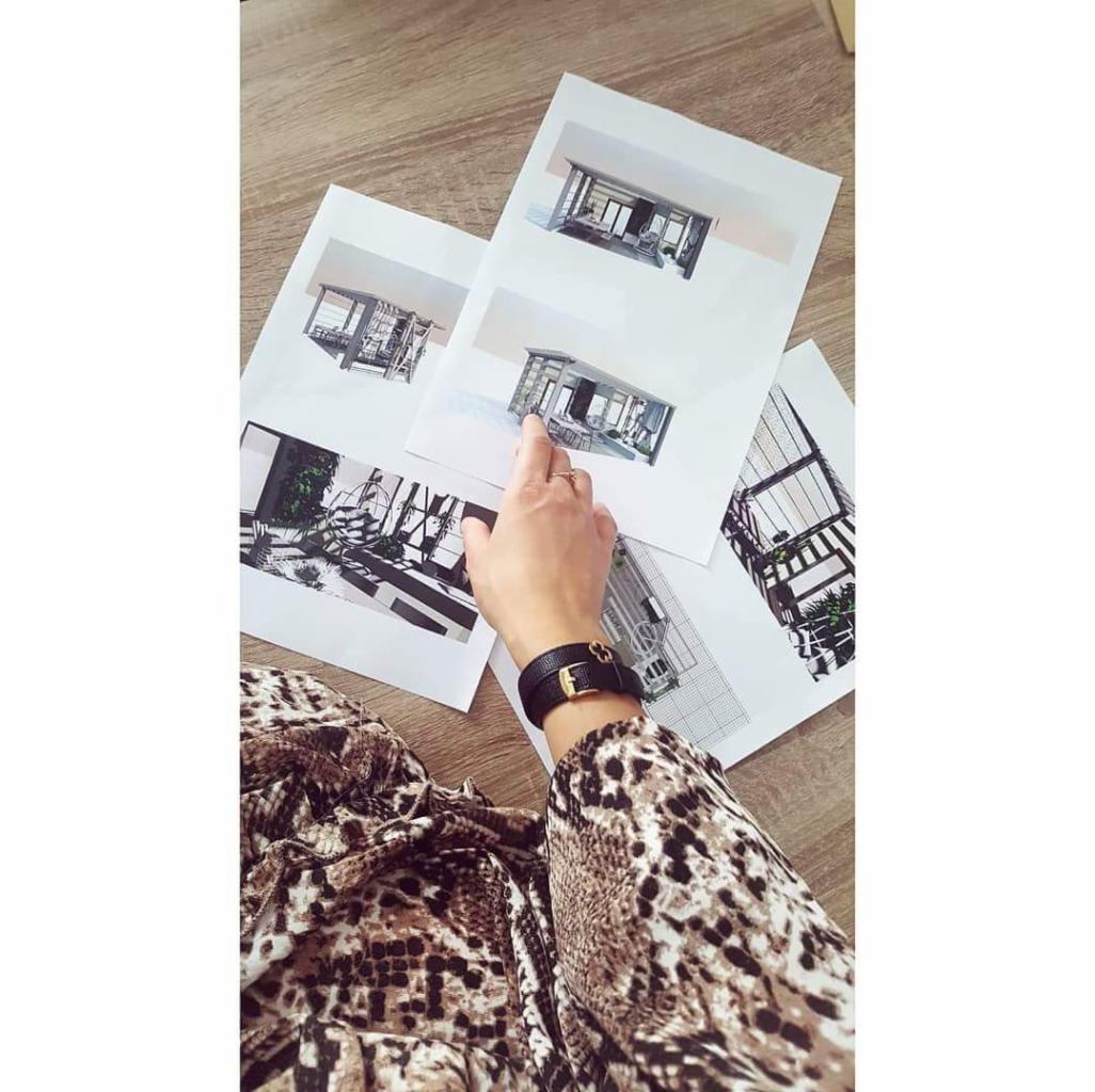 Osoba w bluzce w panterkę wskazuje palcem na wydrukowane wizualizacje 3D nowoczesnych domów z dużymi przeszkleniami, leżące na drewnianym stole.