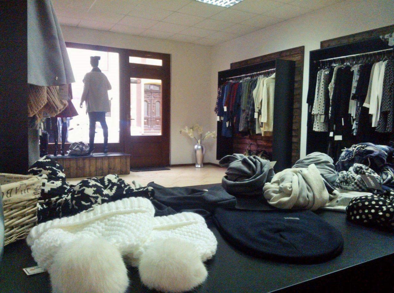 Wnętrze butiku odzieżowego z manekinem w drzwiach, wieszakami z ubraniami oraz stosem czapek i rękawiczek na pierwszym planie.