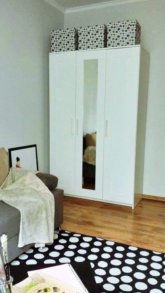 Biała szafa trzydrzwiowa z lustrem w centralnej części, na górze trzy pudełka w czarno-białe wzory, obok szara sofa z narzutą, na podłodze czarno-biały dywan w grochy.