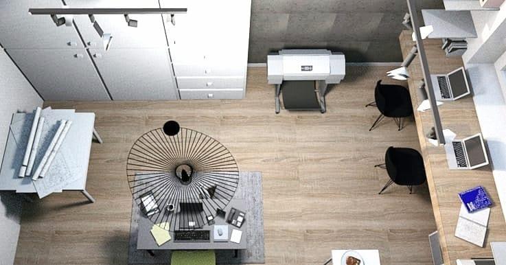 Widok z góry na nowoczesne biuro z drewnianą podłogą, długim biurkiem z laptopami, okrągłym stolikiem z lampą, drukarką i planami na stole w rogu.