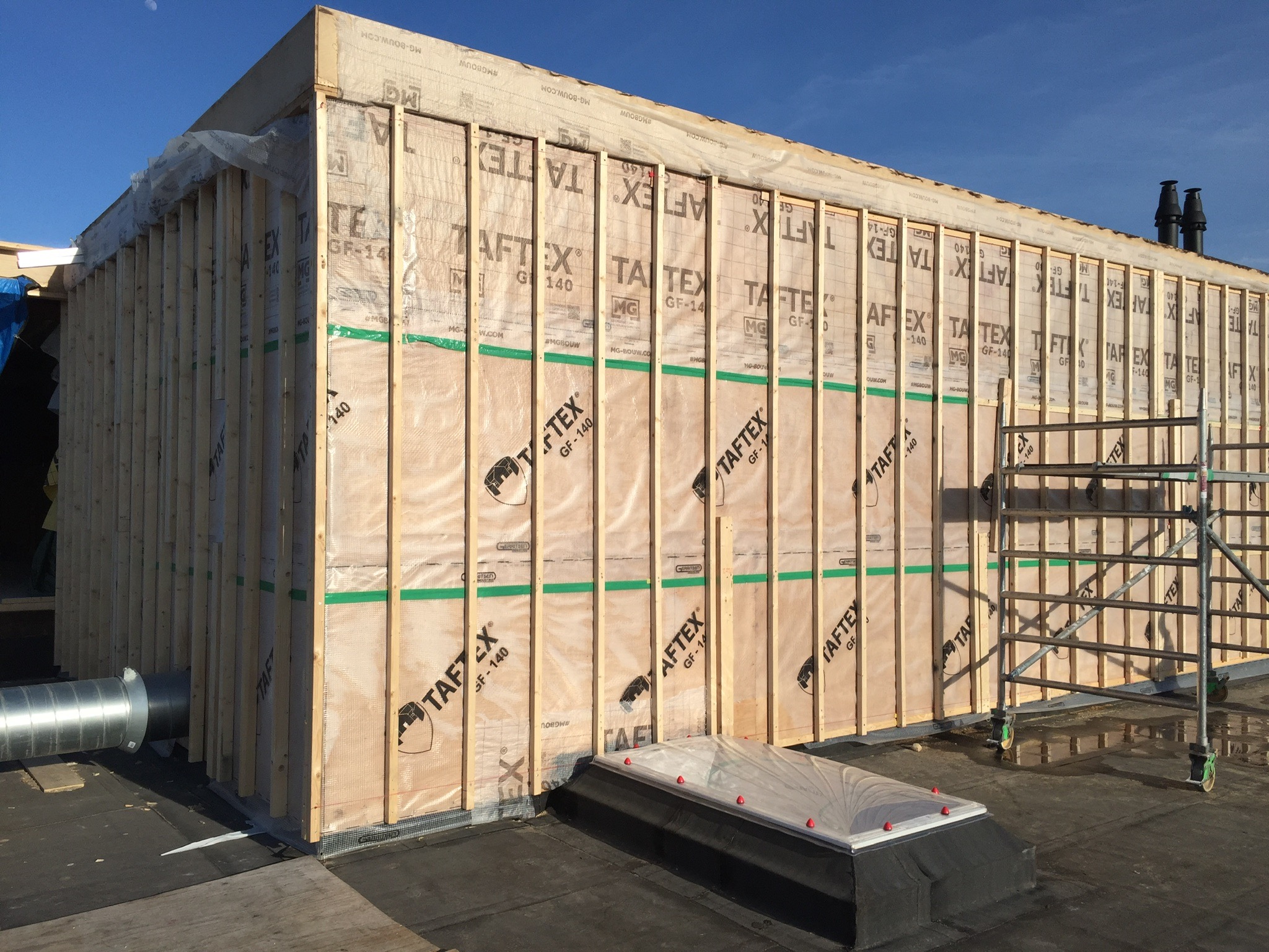 Konstrukcja szkieletowa budynku w trakcie budowy, widoczne drewniane słupki, membrana TAF-TEX, okno dachowe i rusztowanie na dachu.