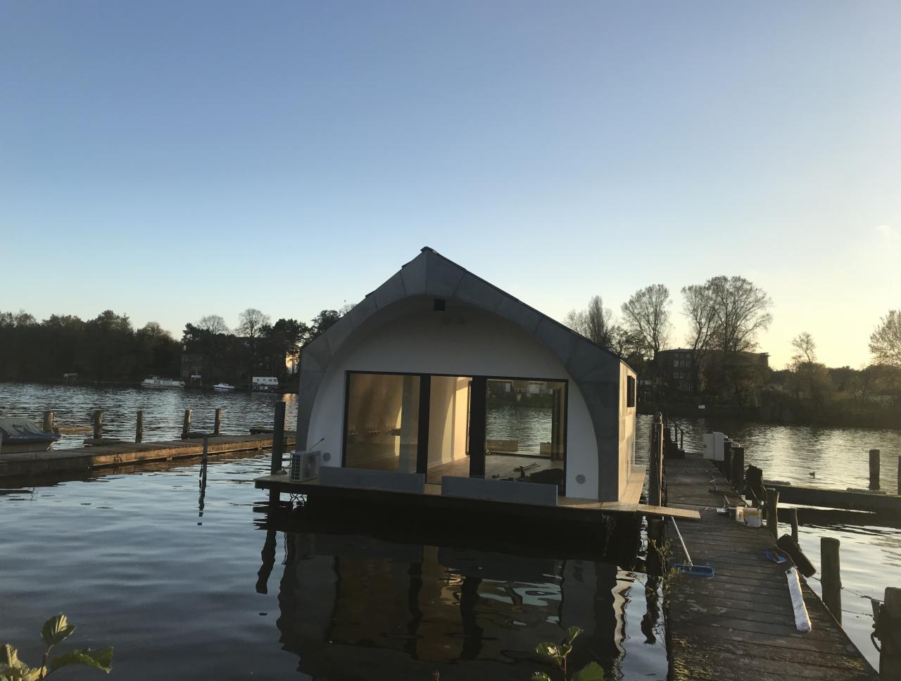 Nowoczesny, pływający dom o nietypowym, łukowym kształcie dachu, umieszczony na drewnianym pomoście nad jeziorem w słoneczny dzień.