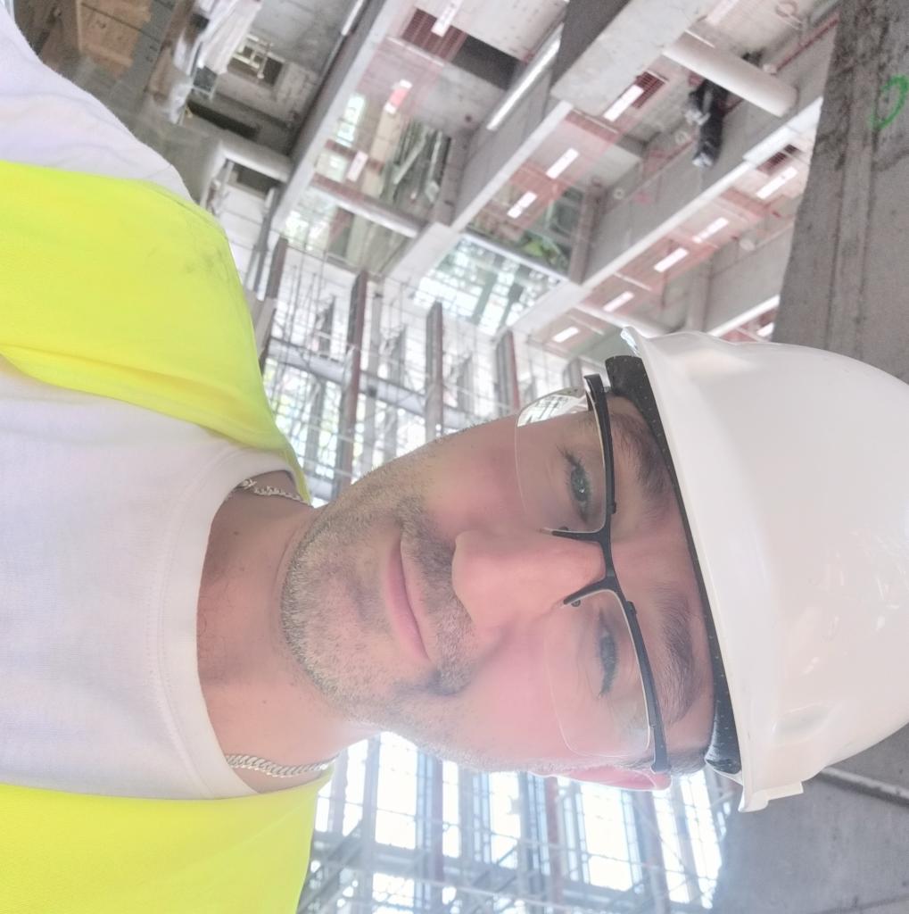 Budowlaniec w kasku i okularach ochronnych na tle surowej konstrukcji budynku w trakcie budowy, widok z dołu.