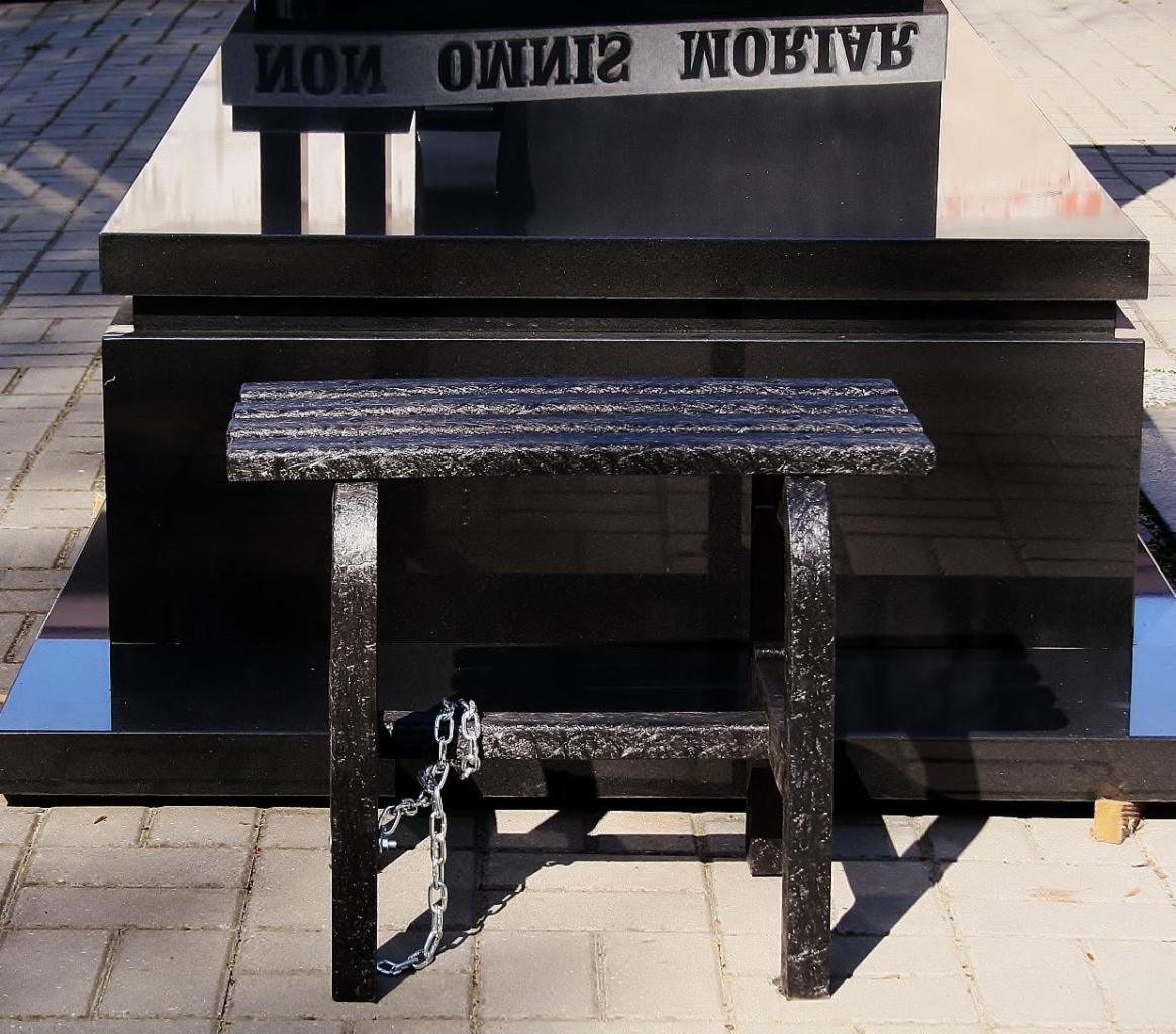 Ławka cmentarna z recyklatu tworzyw sztucznych, z metalowym łańcuchem zwisającym z boku, ustawiona przed czarnym, błyszczącym nagrobkiem.