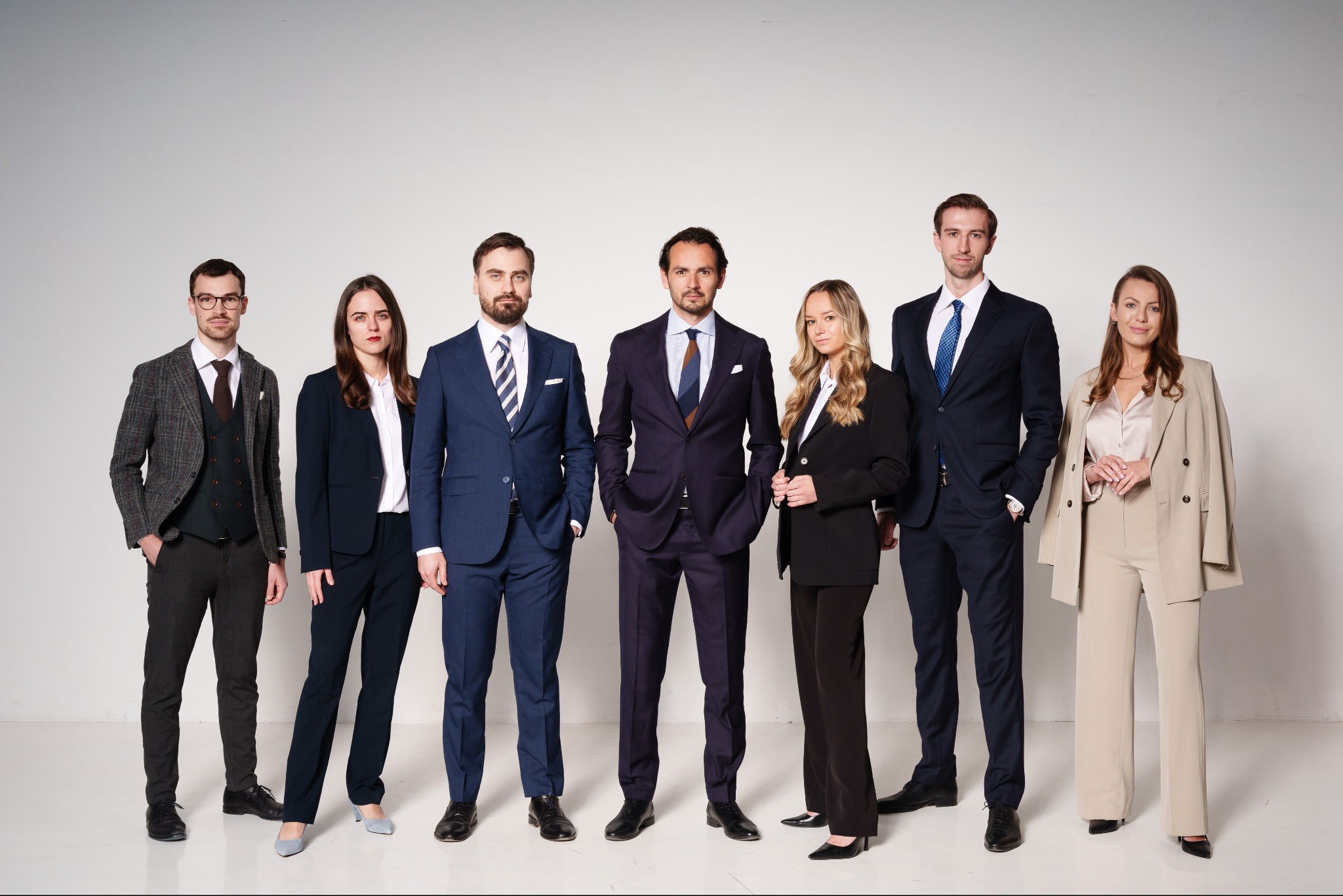 Zespół prawników w garniturach, stojący na białym tle. Różnorodność stylów i kolorów garniturów, od tweedowych po klasyczne granatowe i beżowe, tworzy profesjonalny wizerunek.