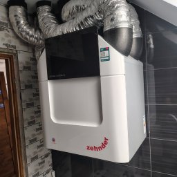 LECH KRUPIŃSKI - Biała jednostka rekuperacyjna Zehnder ComfoAir Q z podłączonymi przewodami wentylacyjnymi, zamontowana w pomieszczeniu z ciemnymi płytkami. Widoczne detale instalacji.